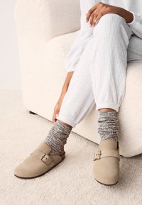 Chaussures beige en daim à enfiler avec un détail boucle, portées avec des chaussettes grises texturées en maille, posées sur un tapis clair près d'un canapé crème.
