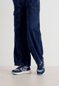 Kék farmer cargo nadrág széles lábakkal és oldalsó zsebekkel, sportos sötétkék és világoskék sneakerrel, fehér kiegészítőkkel párosítva.