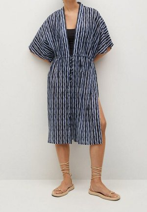 Frau trägt ein knielanges Kimonokleid in Marineblau mit weißen vertikalen Streifen, weitem Ärmel und beigefarbenen, flachen Sandalen mit Schnürung.