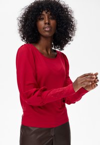 Kobieta z kręconymi włosami, ubrana w czerwony top z długim rękawem i haftowanymi mankietami oraz ciemnobrązowe skórzane spodnie, na jednolitym białym tle.