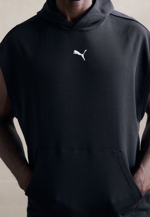 Person iført sort ærmeløs hættetrøje med hvidt Puma-logo midt på brystet og hænder i forlommen.