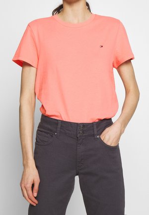 Koraalrood katoenen T-shirt met korte mouwen en een ronde hals, met een klein logo op de linkerborst. Gecombineerd met donkergrijze jeans.