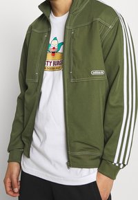 Grön zip-up jacka med hög krage, vita ränder på ärmarna och vit sömnad. Bärs över en grafisk vit t-shirt.