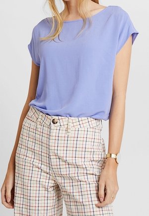 Kobieta ubrana w luźny, liliowy top z krótkim rękawem, wpuszczony do wysokich beżowych spodni w kratę z guzikiem i szlufkami na pasek.