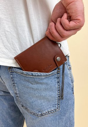 Portafoglio in pelle marrone parzialmente dentro una tasca di denim blu chiaro, dotato di un pulsante a scatto argentato e di una catena attaccata per un trasporto sicuro.