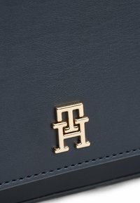 Ciemnoniebieska skórzana torebka z wyraźnym złotym logo monogramu i finezyjnymi stębnowaniami wzdłuż krawędzi. Gładka tekstura z błyszczącym wykończeniem.