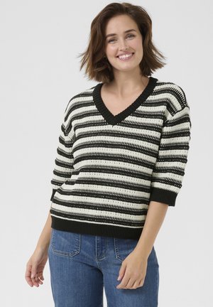 Striped sweater in black and cream, featuring a V-neck and short sleeves. Ribbed hem and cuffs. Worn with blue jeans.