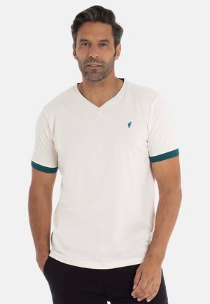 Homme aux cheveux noirs et à la courte barbe portant un t-shirt blanc à col en V avec des bordures turquoise sur les manches et un logo turquoise, un pantalon noir, se tenant devant un fond blanc.