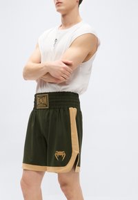 Zöld sportnadrág bézs szegéllyel, rugalmas derékrésszel és arany Venum logóval. Könnyű anyagból, térdig érő, oldalsó hasítékokkal.