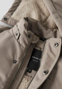 Beige yderjakke med fleece-fôr, lynlåslukning og trykknapper. Mærket viser "name it" på en sort baggrund.