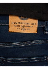 Tmavě modré džíny mají hnědou koženou nášivku s vyraženým textem "Denim Driven Since 1989", připevněnou na zadním pásku.