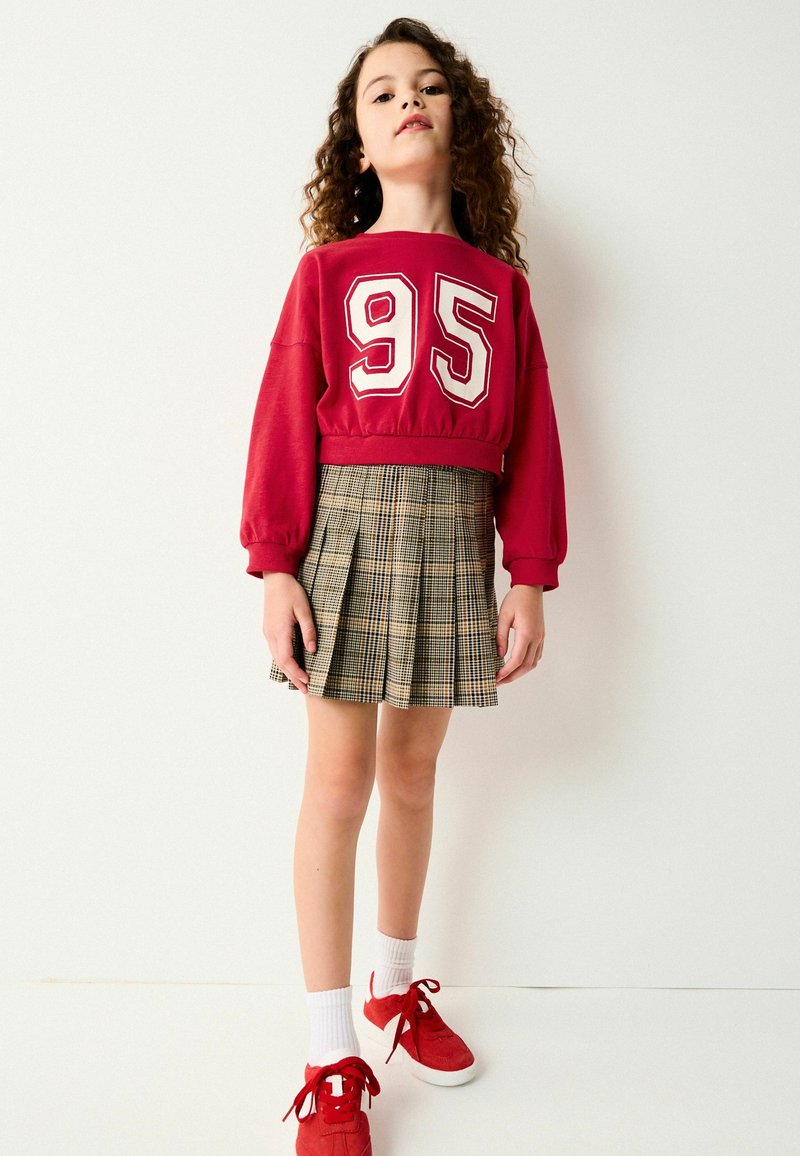 Rød cropped sweatshirt med hvid "95" print, parret med en ternet A-line nederdel. Modellen har røde sneakers og hvide ankelsokker på.