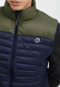 Paminkštintas liemenė su alyvuogių žalios spalvos viršutine dalimi ir tamsiai mėlyna apatine dalimi. Yra aukštas apykaklė, priekinis užtrauktukas ir apvalus logotipo emblema.