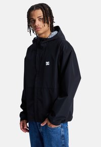 Veste imperméable noire avec capuche, arborant un logo blanc sur la poitrine et une doublure à motifs. Tissu texturé et coupe décontractée.