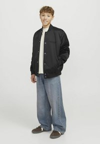 Svart satin bomberjacka med knappar, ribbade muddar och broderad logotyp. Bärs över en ljus skjorta och i kombination med löst sittande ljus denimjeans.