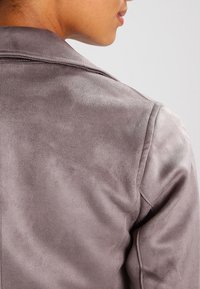 Veste en faux suède gris avec une texture lisse, un col châle et un design ajusté. Un gros plan de l'arrière montre les détails de couture et les contours.