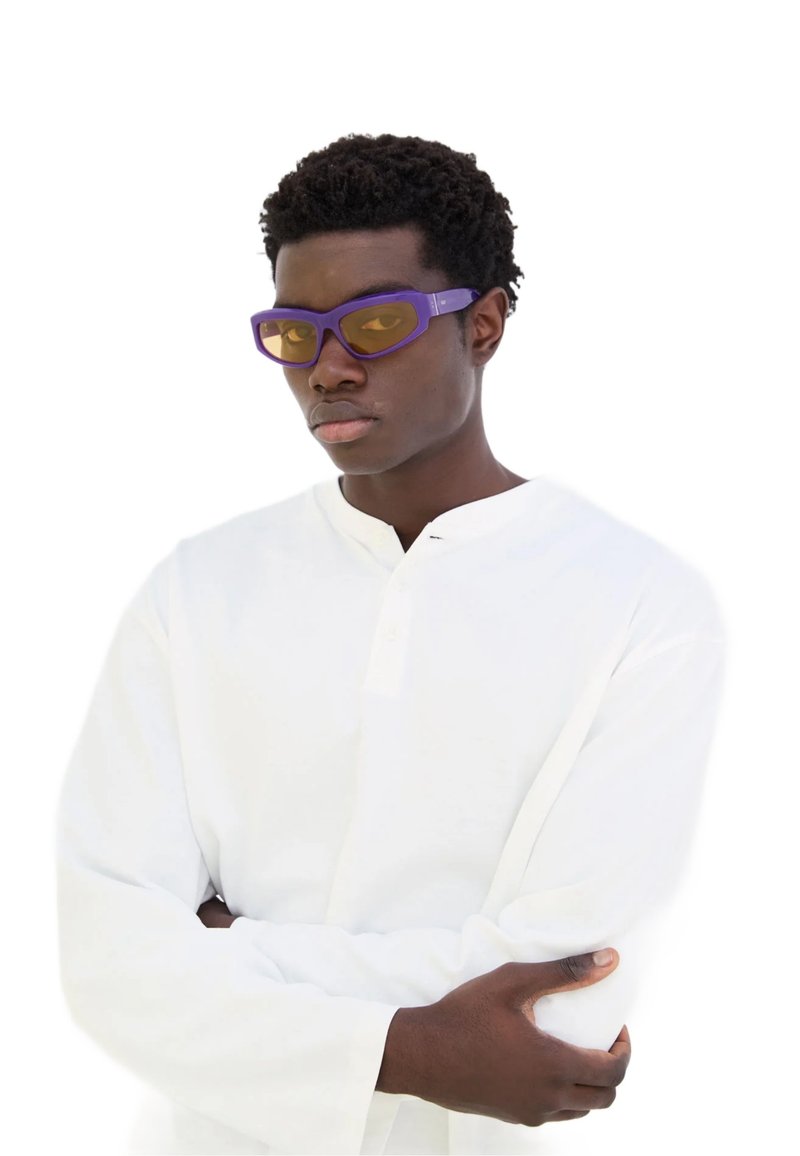 Occhiali da sole con montatura viola e lenti gialle, dal design sportivo. Il modello indossa una maglietta bianca a maniche lunghe con colletto a bottoni.