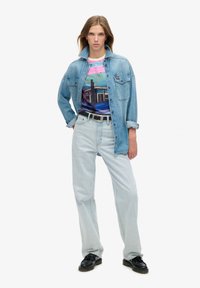 Chaqueta de mezclilla sobre una camiseta gráfica colorida con un paisaje. Jeans azul claro y mocasines negros completan el conjunto.