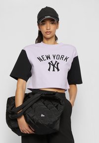 T-shirt cropped in lavanda e nero con stampa "NEW YORK". Tiene in mano una borsa nera trapuntata con doppio manico e dettaglio del logo.