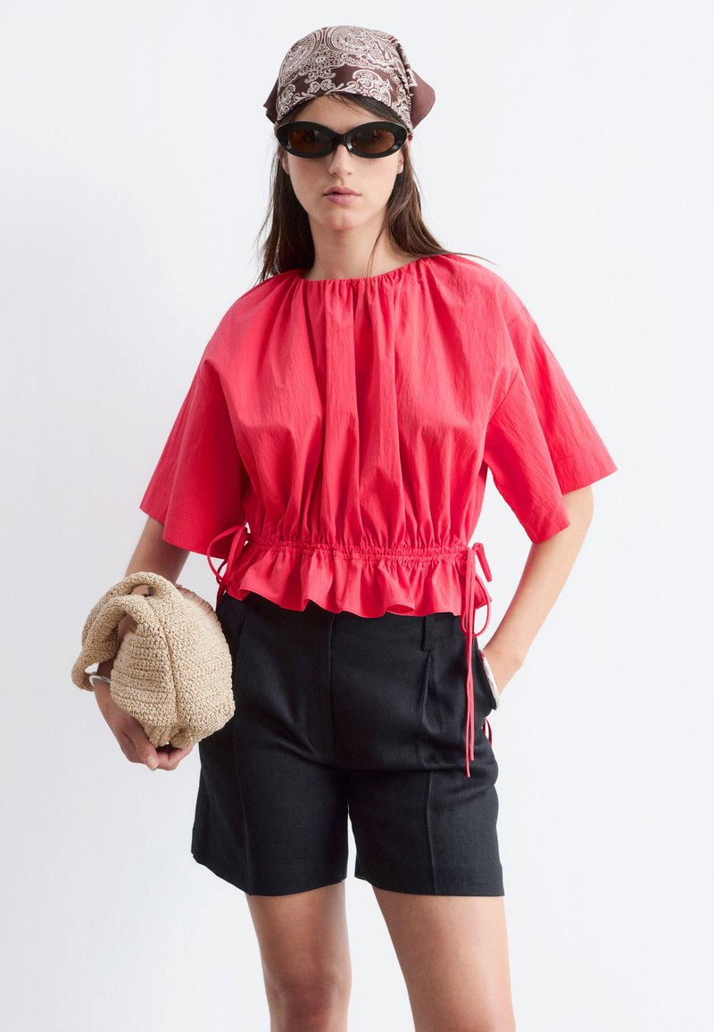 Femme portant un chemisier rouge froncé à la taille, un short noir, un foulard marron à motifs sur la tête, des lunettes de soleil noires, tenant un sac tressé beige, posant sur fond blanc.
