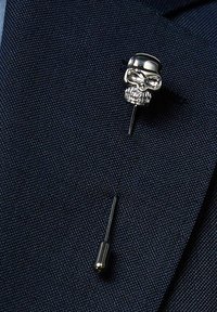 Hopeapintainen pääkallon solki, jossa on yksityiskohtaiset kasvonpiirteet, kiinnitetty ohueen mustaan nuppineulaan, tumman teksturoidun kankaan taustalla.