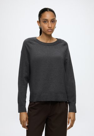 Femme aux cheveux tirés en arrière porte un pull large gris foncé et un pantalon marron foncé, debout devant un fond blanc uni.