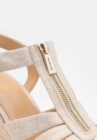 Sandaļa de cuña beige que presenta un upper de tela texturizada, detalle de cremallera dorada y una plantilla de cuero suave. Diseño de tiras para mayor soporte.