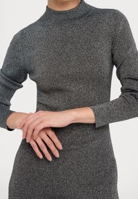 Pull gris ajusté avec un col haut et des manches longues, fabriqué dans un matériau texturé et scintillant. Les mains sont doucement croisées devant.