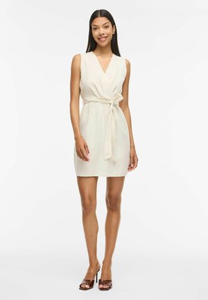 Femme debout souriante portant une robe portefeuille beige sans manches avec ceinture nouée et des talons ouverts marron sur fond blanc.