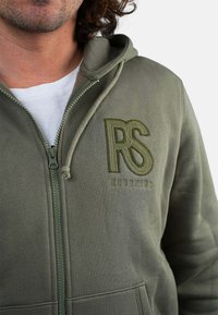 Homme portant un hoodie vert olive avec zip, cordons de serrage et logo "RS REDSKINS" brodé sur la poitrine, par-dessus un t-shirt blanc.