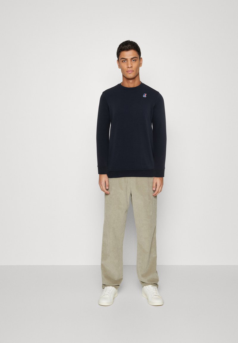 Navy blue long-sleeve sweatshirt with ribbed cuffs and hem, featuring a small logo on the chest, paired with light beige corduroy trousers.