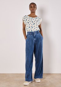Haut blanc avec motif d'oiseaux bleus, associé à un pantalon en denim large taille haute, bleu clair. Le modèle porte des baskets crème. Fond simple.