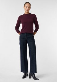 Vrouw die met handen achter haar rug staat, gekleed in een paarse langemouwen trui, donkere wijde jeans met knopen en zwarte enkellaarsjes met hak.