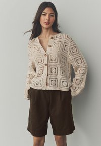 Krämmig stickad cardigan med fyrkantiga mönster, knäppning fram och långa ärmar, ihopparad med mörkbruna shorts. Mjuk, strukturerad utseende.