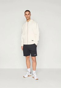 Κρέμα φούτερ με κλείσιμο, μαύρες βερμούδες, λευκά αθλητικά παπούτσια με καφέ λεπτομέρειες. Το μοντέλο στέκεται σε ουδέτερο φόντο, επιδεικνύοντας την εφαρμογή του ρούχου.