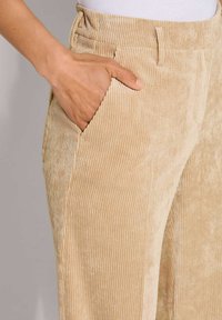 Beige corduroy broek met verticale ribbeltextuur, voorzien van een voorzak en een tailleband. Close-up toont een hand die in de zak rust.