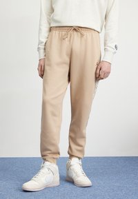 Beige byxor i sweatpants-stil med elastisk midja och dragsko. Sidostrips har ett mönster. Kombineras med vita högskurna sneakers.