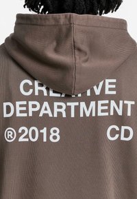 Brown hoodie with a large white print on the back that reads "CREATIVE DEPARTMENT ® 2018 CD." Features a drawstring hood.