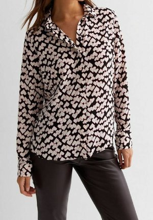 Mujer con camiseta negra de manga larga con estampado de corazones blancos y pantalones de cuero negros, de pie contra un fondo claro.