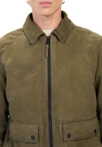 Khaki læderjakke med lynlås foran, spidse kraver, to forlommer og en struktureret overflade. Har et mærke med teksten "ADVENTURES."