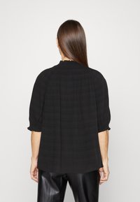Blouse noire à manches bouffantes courtes et encolure froncée. La texture présente un motif à carreaux subtil. Coupe ample, associée à un pantalon noir brillant.