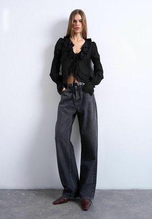 Kvinde iført en sort bluse med flæser, løse mørkegrå jeans med sort bælte og spidse mørkebrune sko, stående foran en hvid væg.