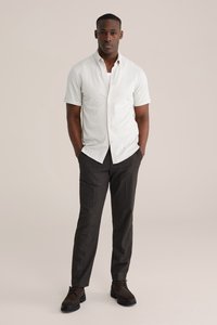 Korte mouw, overhemd met knopen in lichtbeige stof gecombineerd met donkergrijze broek. Schoeisel bestaat uit donkerbruine veterschoenen.