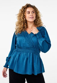 Blouse teal avec un décolleté en V profond et une taille smockée, dotée de manches longues et d'un ourlet peplum fluide. Tissu lisse avec une texture subtile.