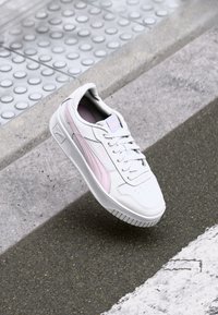 White sneakers with light pink accents, made of smooth leather. Features a perforated toe box, lace-up design, and textured rubber sole.