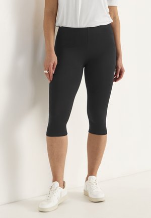 Czarne legginsy capri wykonane z elastycznego materiału, o dopasowanym kroju, sięgające do połowy łydki, zestawione z białymi butami sportowymi.