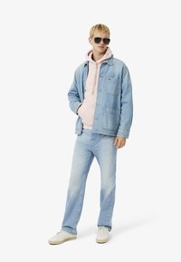 Chaqueta de mezclilla azul claro sobre una sudadera rosa, combinada con jeans azul claro y zapatillas blancas. Los accesorios incluyen gafas de sol oscuras.