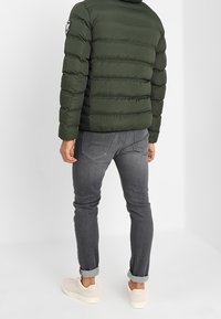 Grön puffjacka med quiltrad design och logotypapplikation på vänster ärm, i kombination med grå jeans och ljusa sneakers.