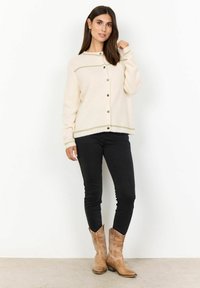 Cream-colored knit cardigan with ribbed texture, button front, and contrasting green stitching along the hem and sleeve cuffs. Worn with black jeans.
