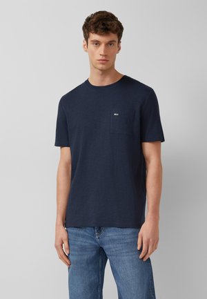 Młody mężczyzna ubrany w granatowy t-shirt z kieszenią i niebieskie dżinsy, stojący na tle jednolitego szarego tła.